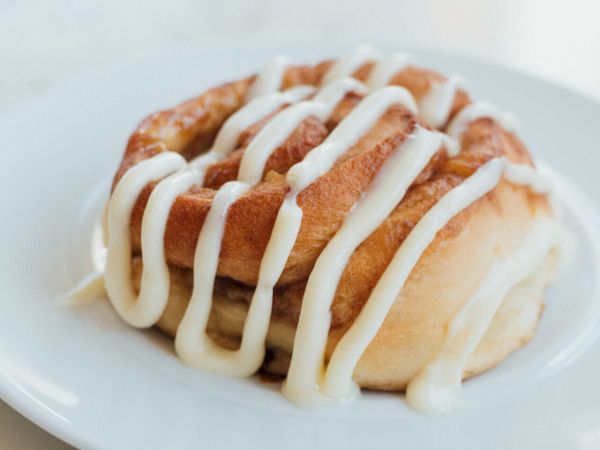A cinnamon roll topped with white icing drizzled in zigzags sits on a white plate, served on a marble table.