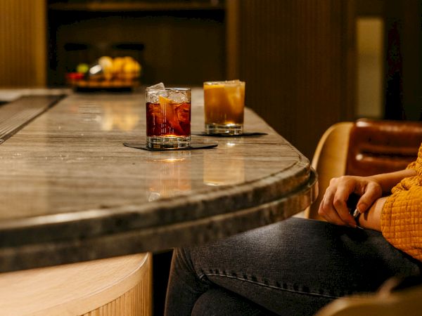 Two drinks sit on a marble bar while a person in a yellow sweater relaxes nearby, legs crossed, in a cozy lounge setup.